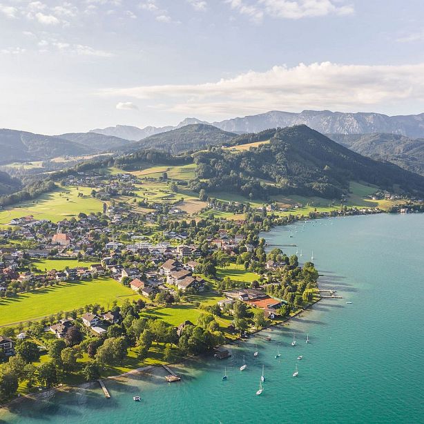 Aerial view of a village by a lake with green hills and distant mountains under a partly cloudy sky.