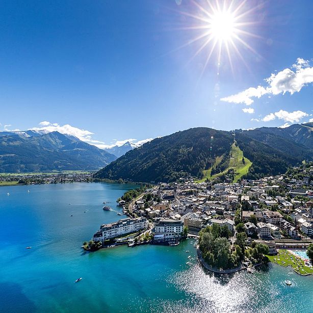 Aerial view of Zell am See, Austria, with a lake, mountains, and a sunny sky. The town is nestled between water and hills, offering scenic natural beauty.