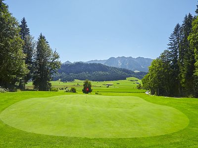 Ein gepflegtes Golfgrün inmitten einer idyllischen Landschaft mit Bergen im Hintergrund. Der Himmel ist klar und die Sonne scheint hell.