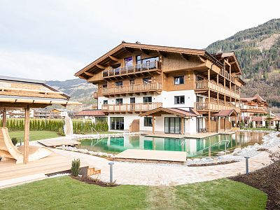 Alpine-style lodge with wooden facade, surrounded by mountains and a modern pool, under a cloudy sky.