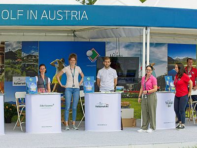 People at a promotional booth labeled 'Golf in Austria,' with scenic background images. They stand behind counters displaying brochures and smiling at the camera.