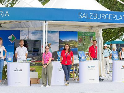 Ein Messestand mit dem Thema "SalzburgerLand" zeigt Menschen hinter Infotischen. Im Hintergrund sind Landschaftsbilder und ein Zelt mit blauem Banner sichtbar.