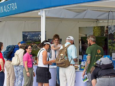 People gather at an outdoor tourist booth in Austria, engaging with staff and collecting travel brochures. The booth is lively with banners and materials displayed.