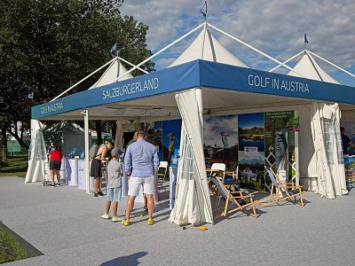 A promotional tent at a golf event with 'Golf in Austria' signage, deck chairs, brochures, and people interacting under a blue sky.