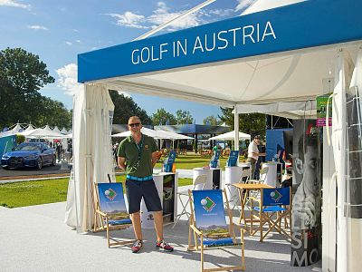 Man in green shirt stands at a booth labeled "Golf in Austria," surrounded by chairs and promotional materials at an outdoor event under a clear blue sky.