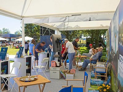 Outdoor booth at a fair with people engaging in various activities. Displays and chairs are set up under a white tent, surrounded by greenery.