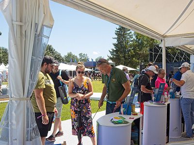 People at an outdoor event, mingling around stalls under tents. It's sunny with trees and grass visible in the background.