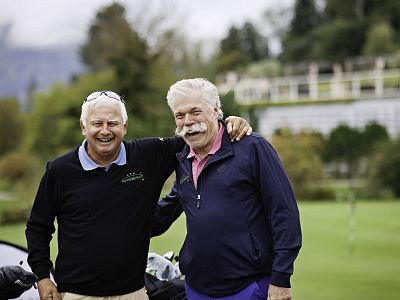 Two older men stand on a golf course smiling, dressed in golf attire. Greenery and a cloudy sky form the backdrop, suggesting a pleasant day outdoors.