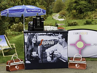 Outdoor coffee stand with Segafredo branding, coffee machine, pastries, umbrella, and banners on a sunny day, set in a park environment.