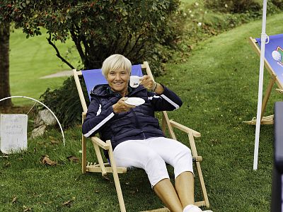 Person lounging in a deck chair on grass, sipping a drink and smiling, with trees and greenery in the background, creating a relaxed outdoor atmosphere.