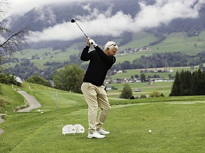 A golfer swings on a green course, surrounded by mountains and clouds. The landscape is lush and vast, creating a serene outdoor setting.