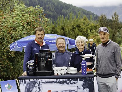 Four people stand together at an outdoor refreshment station with coffee and snacks, surrounded by a lush, green landscape under a cloudy sky.