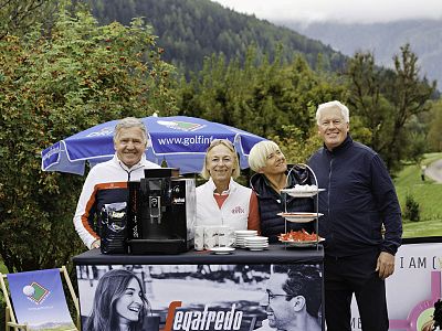 Four people smile in front of a coffee station at a golf course, with trees and mountains in the background, under a cloudy sky.