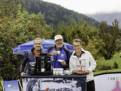 Three people at a small outdoor coffee stand in a lush, mountainous area. They are smiling and holding cups, with greenery and hills in the background.