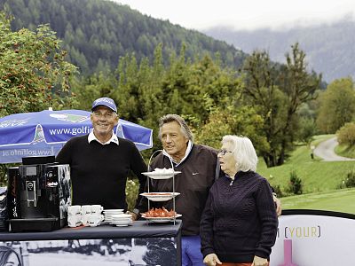 Three people stand by a refreshment table with coffee cups and pastries at a golf course, surrounded by trees and a mountainous landscape in the background.