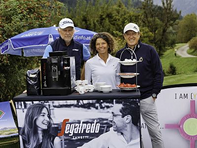 Three people stand smiling behind a coffee station outdoors, with a coffee machine and cups, surrounded by greenery and a promotional umbrella.