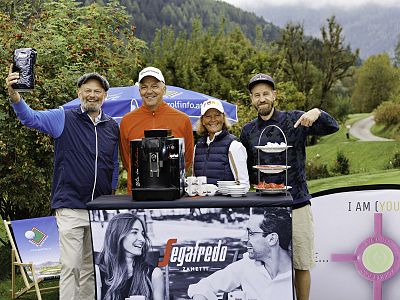 Four people stand behind a table with a coffee machine outdoors. They're smiling, surrounded by trees, with mountains and clouds in the background.