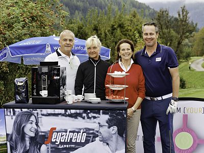 Vier Personen stehen vor einem Kaffee-Stand im Freien, umgeben von grüner Landschaft. Eine Kaffeemaschine und Produkte sind auf einem Tisch vor ihnen platziert.