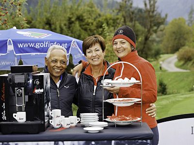 Three people pose happily beside a coffee machine and dessert stand at an outdoor event, with a golf course and trees in the background.