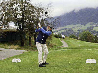 A golfer in blue jacket and beige pants swings on a green course with mountains in the background and cloudy sky overhead.