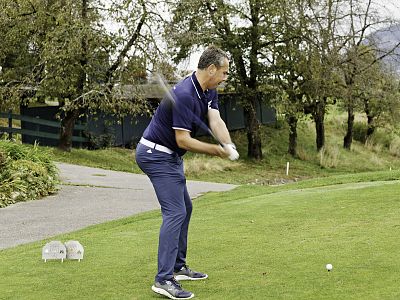 A man in a navy outfit swings a golf club on a lush green course, with trees and a building in the background.