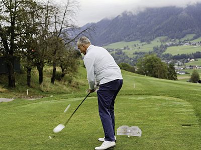 Man playing golf on a course with mountains and cloudy sky in background; mid-swing with lush scenery around.