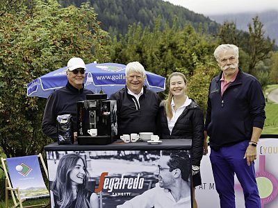 Four people stand smiling outdoors by a promotional stand, with greenery and a cloudy sky behind them, dressed in mixed casual and sporty attire.