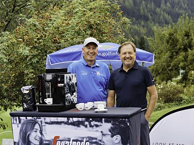 Two men stand beside a coffee setup at a golf event, surrounded by lush greenery and promotional banners. Both are smiling and the scene has a relaxed ambiance.