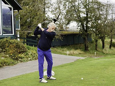 A senior golfer in a dark sweater and blue pants swings a golf club on a green course with trees and a clubhouse in the background.