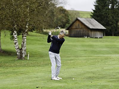 Golfer in mid-swing on a green fairway, wearing a navy sweater and light pants, with trees and a wooden shed in the background under a cloudy sky.