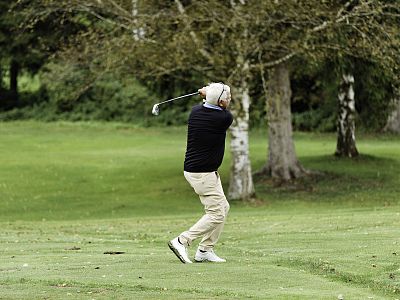 A golfer in mid-swing on a green course, wearing a jacket and pants. Trees are visible in the background, creating a peaceful outdoor environment.