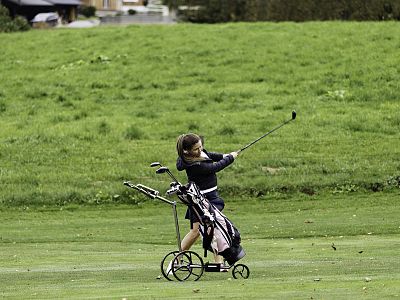 A small child playing golf on a green field, swinging a club with a trolley next to them holding golf equipment.