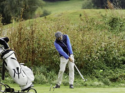 A golfer in a blue jacket swings his golf club on a green course. A golf bag is nearby. Trees and bushes form a lush backdrop.