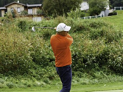 Golfer in an orange shirt taking a swing on a golf course, surrounded by greenery, with houses in the background.