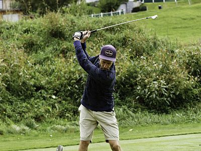 A golfer swings his club on a green course, wearing a cap, dark shirt, and shorts. Dense greenery surrounds the area.