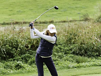 A golfer swings a club on a lush green course, wearing a white cap, navy vest, and blue pants, surrounded by green foliage and grass.