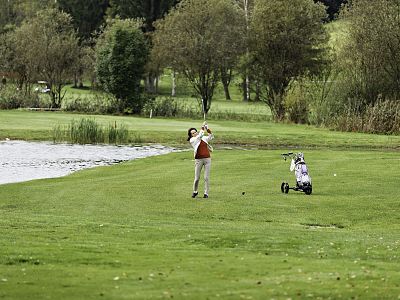A person playing golf swings their club on a green field, with a lake and trees in the background and a golf cart to the side.