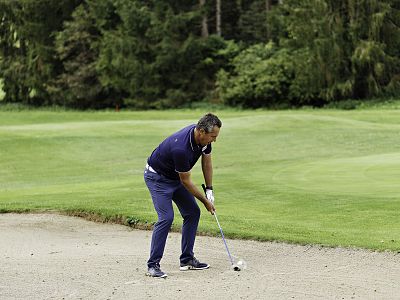A person playing golf in a sand trap, focusing on their shot, surrounded by a green course with tall trees in the background.