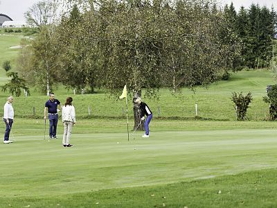 Four people on a golf course, one about to putt, surrounded by green grass and trees under a cloudy sky.
