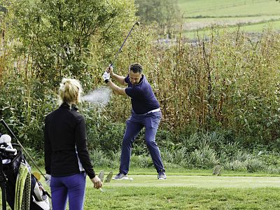 A man swings a golf club on a green course, while a woman watches nearby with a golf bag in the foreground. Trees and fields are in the background.