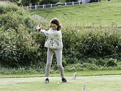 A young girl in a floral jacket and beige pants is swinging a golf club on a green course. Trees and plants are visible in the background under an overcast sky.
