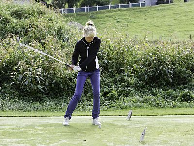 A golfer in a black jacket and blue pants swings a golf club on a lush green course surrounded by bushes and hills.