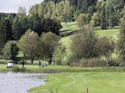 Golfers on a lush green course near a lake, surrounded by trees and hills under a cloudy sky.