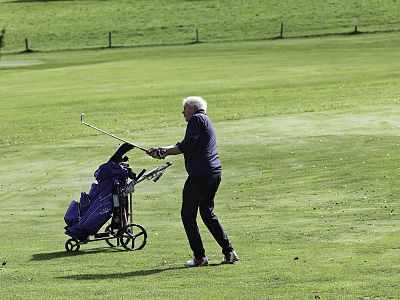 Elderly man golfing on a green course, mid-swing, with a golf bag on a cart beside him on a sunny day.