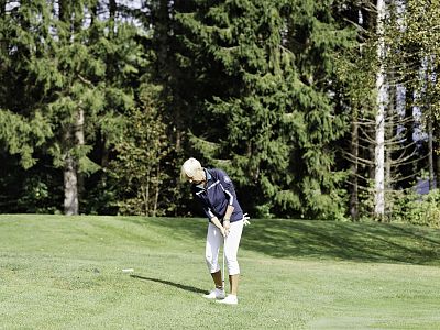 A woman in a navy shirt and white pants plays golf on a grassy course surrounded by tall, dense trees of a forest.