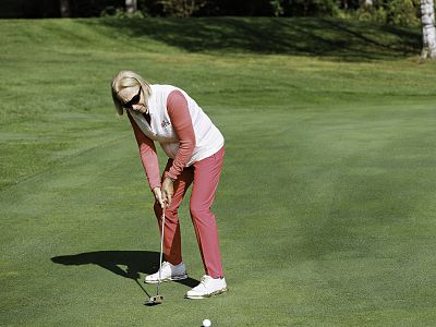 Elderly woman in pink and white attire putts on a sunny green golf course, with trees in the background. She appears focused and is wearing sunglasses.
