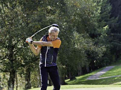 A man with gray hair swings a golf club on a green golf course, surrounded by trees in bright daylight.