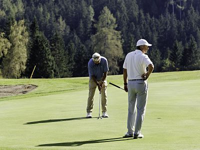 Two men on a golf course; one putting, another watching. Lush green fairway and trees in the background. They wear golf attire, with caps and clubs.
