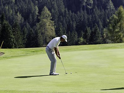 A golfer wearing a white shirt and hat is putting on a green golf course with a forest in the background, under a clear blue sky.