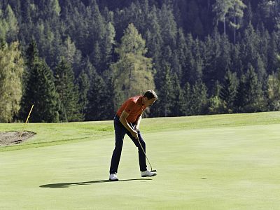 A golfer in mid-swing on a green golf course surrounded by dense forest, concentrating on the ball, under a clear blue sky.